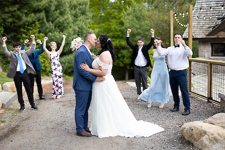 A newlywed couple kisses, surrounded by cheering friends in a celebratory outdoor setting.