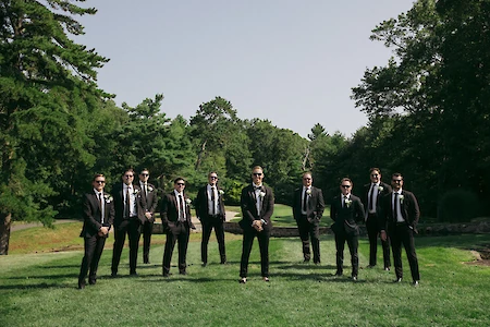 A group of people in suits and sunglasses stand on grass with trees in the background, posing for a formal photo.