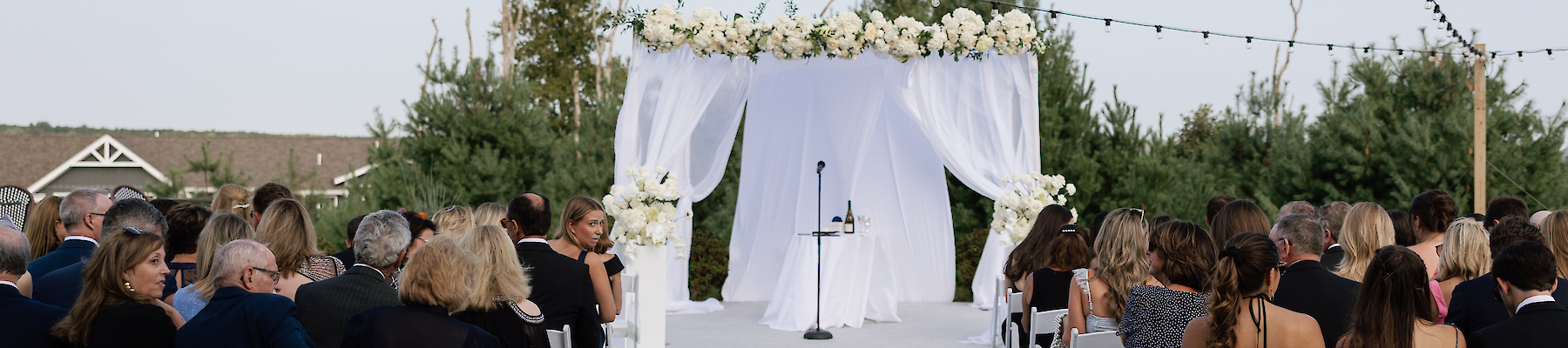 An outdoor wedding setup with a floral-adorned altar, chairs filled with guests, and string lights overhead on a clear day.