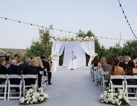 An outdoor wedding setup with a floral-adorned altar, chairs filled with guests, and string lights overhead on a clear day.