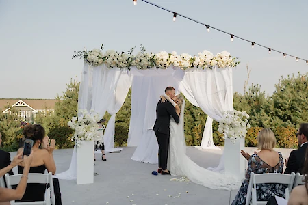 A couple kisses under a floral wedding arch, surrounded by guests seated on white chairs outdoors.