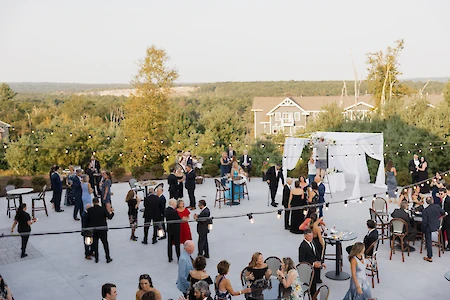 People are gathered outdoors at an event, with a decorated arch, tables, and string lights, set against a scenic backdrop.