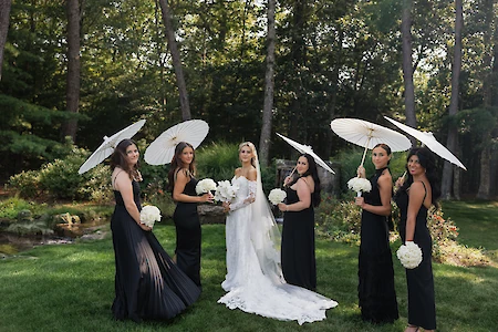 A bride and bridesmaids in black dresses hold white parasols and bouquets, posing outdoors in a wooded area.