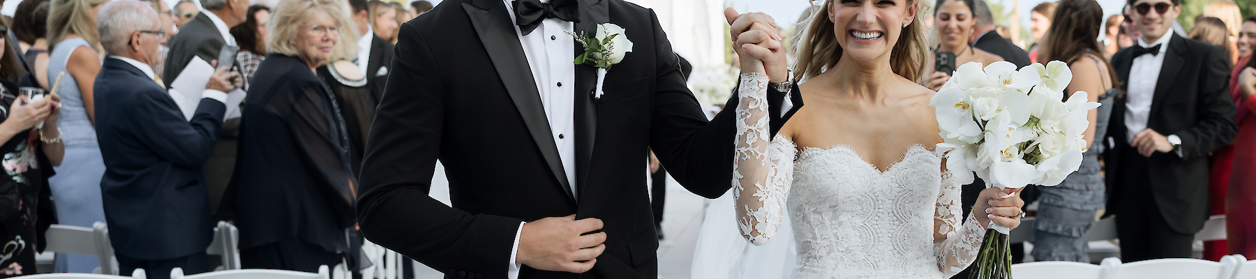 A bride and groom are joyfully walking down an aisle at their wedding, surrounded by guests seated on white chairs.