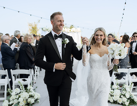 A bride and groom are joyfully walking down an aisle at their wedding, surrounded by guests seated on white chairs.