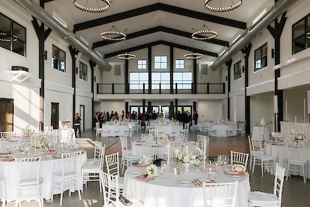 The image shows an elegant banquet hall setup with round tables, white chairs, floral centerpieces, and chandeliers in a spacious venue.