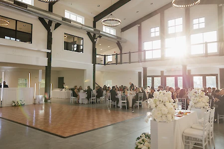 A spacious event hall with guests sitting at decorated tables, featuring a dance floor and flowers, illuminated by sunlight.