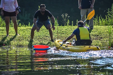 A person in a yellow kayak paddles towards the shore where three people stand watching. A red and blue ring floats nearby.
