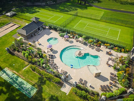 The image shows a luxurious outdoor area with a pool, hot tub, tennis courts, and seating, surrounded by gardens and a small building.