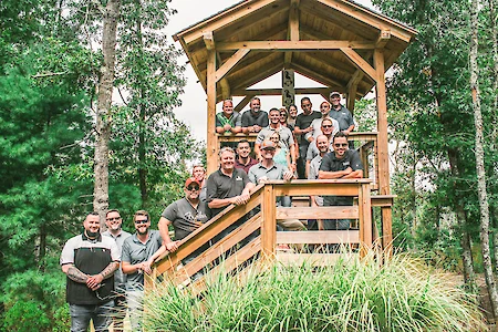 A group of people are gathered on and around a wooden gazebo-like structure in a forested area, smiling and posing for a group photo.
