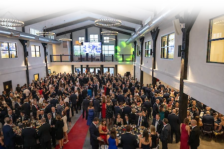 A large gathering in a banquet hall, with people in formal attire socializing and dining at tables under elegant lighting fixtures.