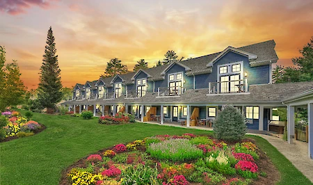 A row of blue two-story houses with porches is set against a sunset backdrop, surrounded by lawns and colorful flower gardens.