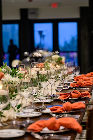 A long dining table set for an event with orange napkins, floral arrangements, and glasses, creating an elegant atmosphere.