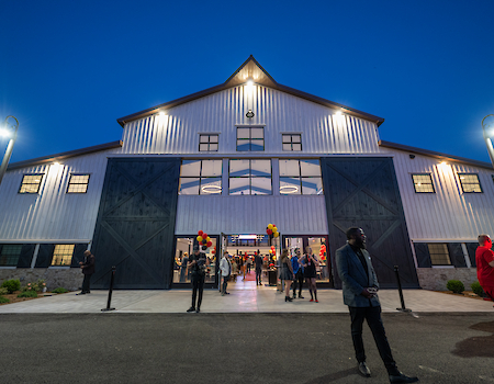 A large barn-style building lit at night with people entering, featuring two big doors, and streetlights illuminating the scene.