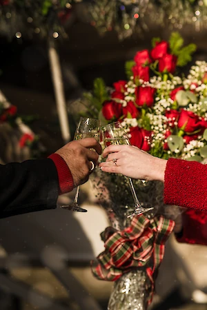Two people clinking champagne glasses in front of a bouquet of red roses, wrapped with a festive bow, creating a celebratory scene.