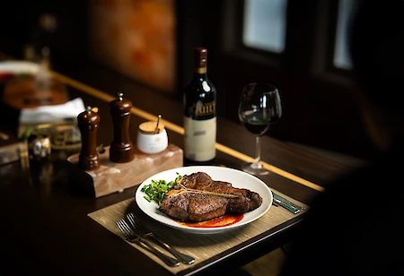A plate with cooked steaks, topped with greens, next to a bottle and glass of red wine, pepper mills, and a small condiment set.