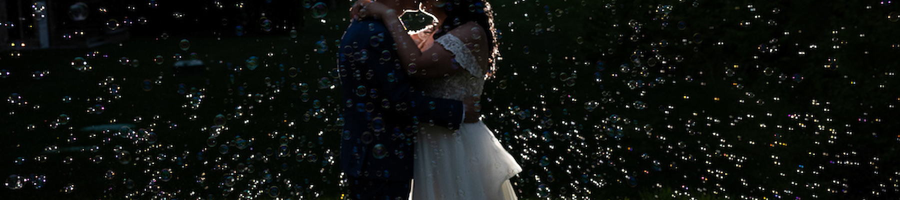 A couple is silhouetted as they embrace among scattered bubbles on a grassy field at dusk.