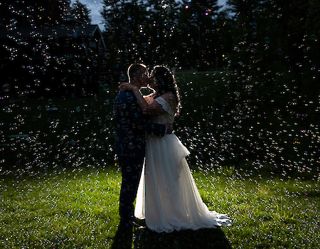 A couple is silhouetted as they embrace among scattered bubbles on a grassy field at dusk.