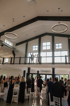 A couple stands on a balcony waving to guests at a gathering in a spacious hall with large windows and elegant lighting fixtures.