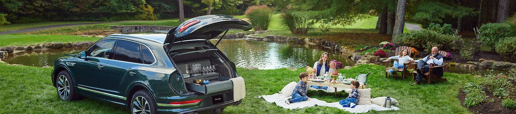 A family is having a picnic on a grassy area near a pond, with an SUV parked nearby.