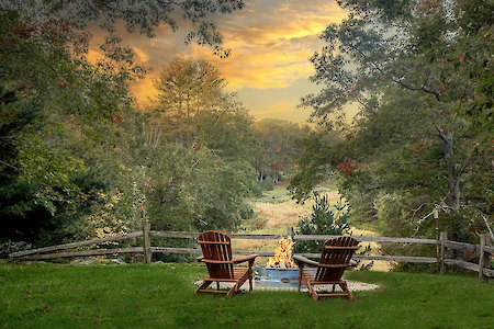 Two chairs face a fire pit on a grassy area, surrounded by trees under a colorful sunset sky, with a wooden fence in the background.