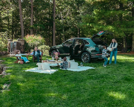 A group enjoying a picnic on grass near a parked SUV, with blankets and pillows, surrounded by trees in the background.