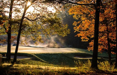 A serene autumn golf course, framed by trees with orange leaves, mist rising in the sunrise light, creating a tranquil scene.