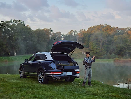 A man in fishing gear stands by a black SUV with an open trunk, by a foggy lakeside surrounded by trees and greenery.
