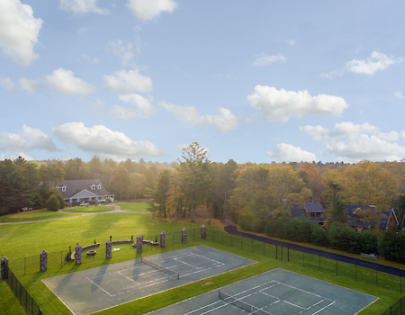 The image shows a scenic landscape featuring tennis courts, surrounded by trees and houses, under a partly cloudy sky.