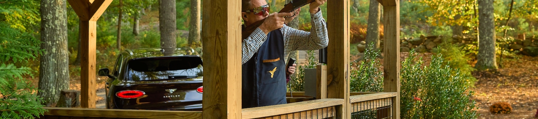 A person is aiming a rifle from a wooden gazebo in a wooded area. There's a black truck parked nearby, and the setting is autumn.