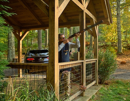A person is aiming a rifle from a wooden gazebo in a wooded area. There's a black truck parked nearby, and the setting is autumn.