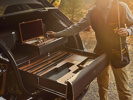 A person is accessing a storage drawer in the trunk of a vehicle, holding items like a case and a rifle, set in an outdoor environment.
