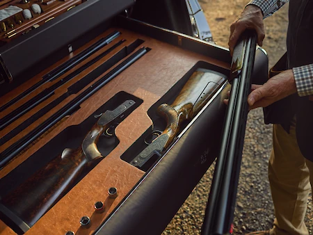 A person is handling a long gun near a storage case with disassembled firearms and ammunition arranged neatly inside.