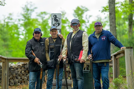 Four people with shotguns pose outside near a wooden fence and sign numbered 12, wearing caps and casual clothing.