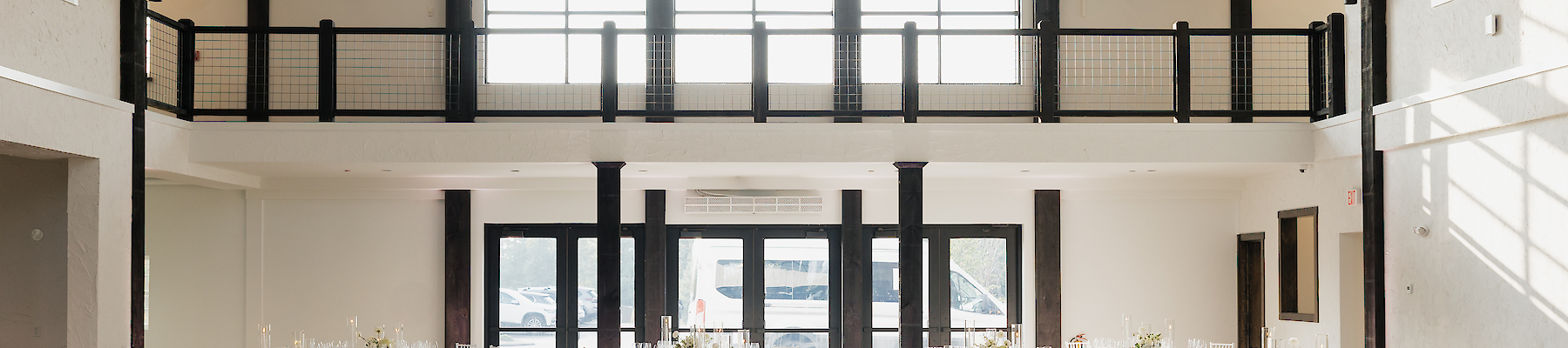 A spacious, elegant venue with white-clothed tables, chairs, and large windows, designed for events or receptions, elegantly lit.