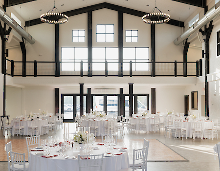 A spacious, elegant venue with white-clothed tables, chairs, and large windows, designed for events or receptions, elegantly lit.