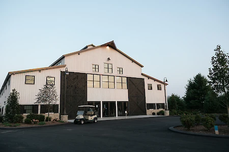 The image shows a large white building with dark wooden accents, large windows, and a golf cart in front, surrounded by greenery.