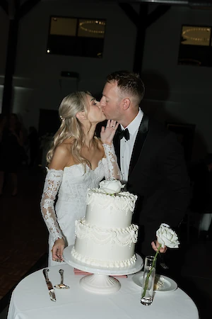 A couple is kissing in front of a white, tiered wedding cake on a table, with a rose in a vase nearby.