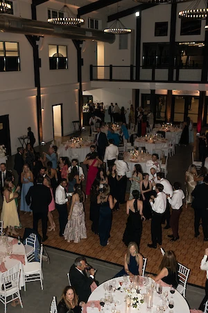 A large indoor event with people gathered around tables and dancing on a wooden floor, in an elegantly decorated venue.