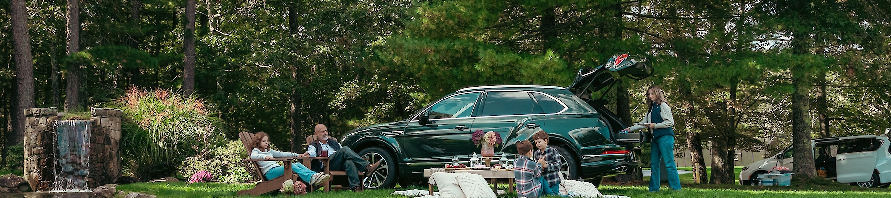 A family enjoys a picnic by a lakeside surrounded by trees with a parked SUV nearby, reflecting a serene outdoor setting.