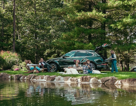 A family enjoys a picnic by a lakeside surrounded by trees with a parked SUV nearby, reflecting a serene outdoor setting.