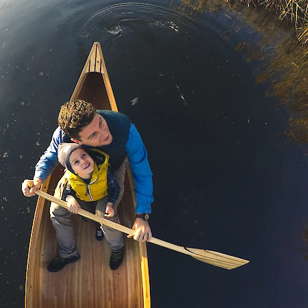 A person and a child are in a canoe on a calm body of water, with the child holding a paddle and both looking upward.