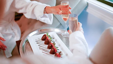 Two people in robes toast with champagne over a plate of chocolate-covered strawberries on a tray.