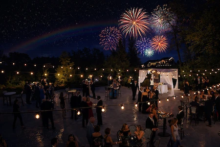 A nighttime outdoor event with people socializing under string lights, fireworks in the sky, and a rainbow visible.