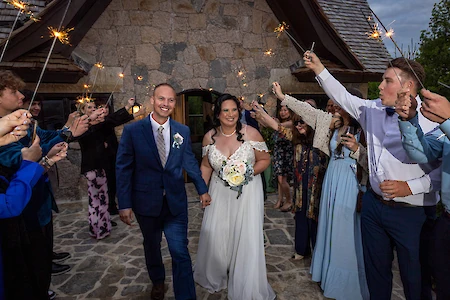A couple is walking through a crowd holding sparklers, in front of a stone building.