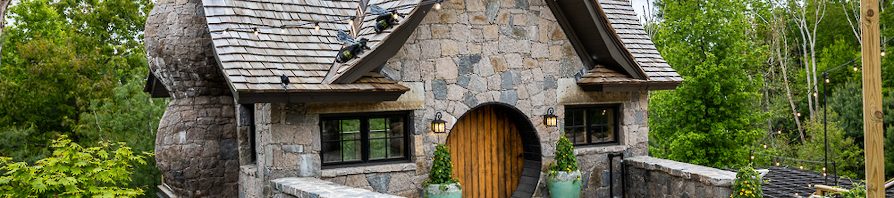 A stone cottage with a curved roof, circular door, and lush greenery surrounds it. Pathway leads to the entrance under cloudy skies.
