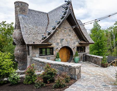 A stone cottage with a curved roof, circular door, and lush greenery surrounds it. Pathway leads to the entrance under cloudy skies.