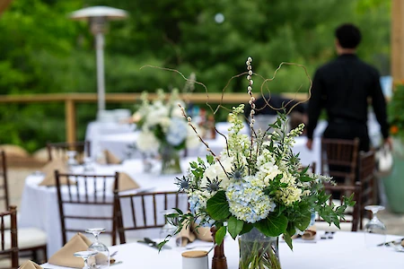 A floral centerpiece adorns a table set with white linens; a person dressed in black stands in the background, outdoors.