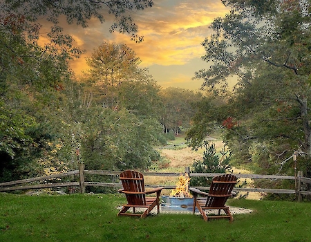 Two chairs face a fire pit on a grassy area, surrounded by trees and a wooden fence, with a scenic view under a colorful, cloudy sky.