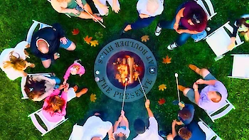 Aerial view of people gathered around a fire pit, roasting marshmallows with sticks, on grass with scattered autumn leaves ending the sentence.